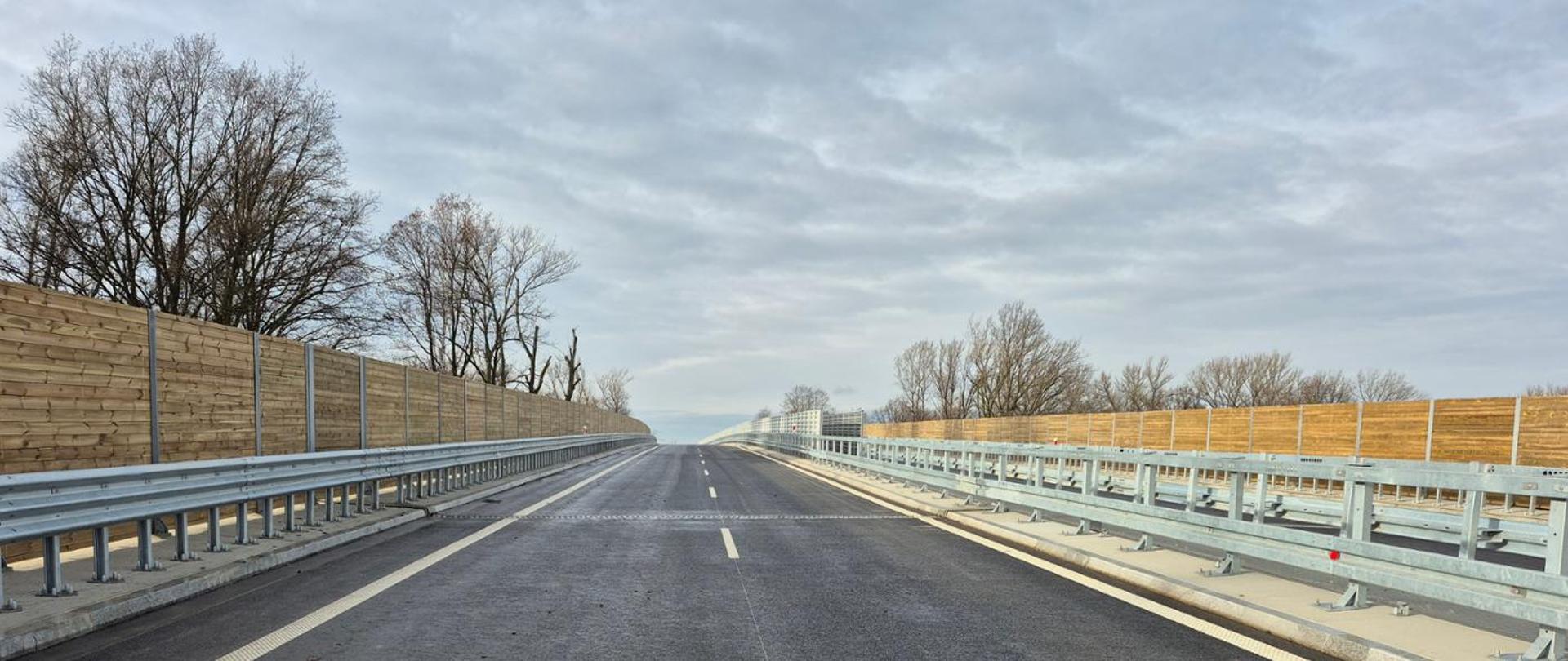 Nowa droga z asfaltową nawierzchnią na wiadukcie, po obu stronach metalowe bariery ochronne i drewniane ekrany akustyczne. W tle widoczne bezlistne drzewa i pochmurne niebo.