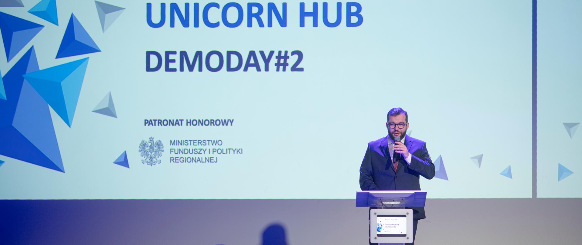Na zdjęciu przy mównicy stoi wiceminister Grzegorz Puda, w tle jasny ekran z napisem Unicorn Hub DemoDay#2, z przodu osoby siedzące na widowni.