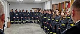Na zdjęciu w sali konferencyjnej Komendy Powiatowej Państwowej Straży Pożarnej widoczni strażacy Państwowej Straży Pożarnej i druhowie kończący szkolenie ubrani w ubrania koszarowe w czarnym kolorze podczas uroczystego apelu kończącego szkolenie.