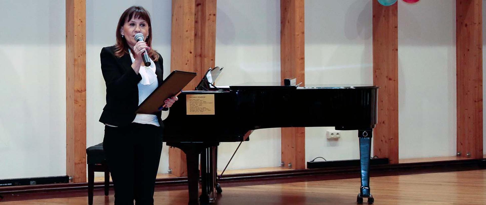 Na zdjęciu w pomieszczeniu, z lewej strony stoi kobieta zwrócona twarzą do widza, w lewej ręce trzyma teczkę, w prawej mikrofon przez który coś mówi. Z tyłu przy biało - brązowej ścianie stoi czarny fortepian.