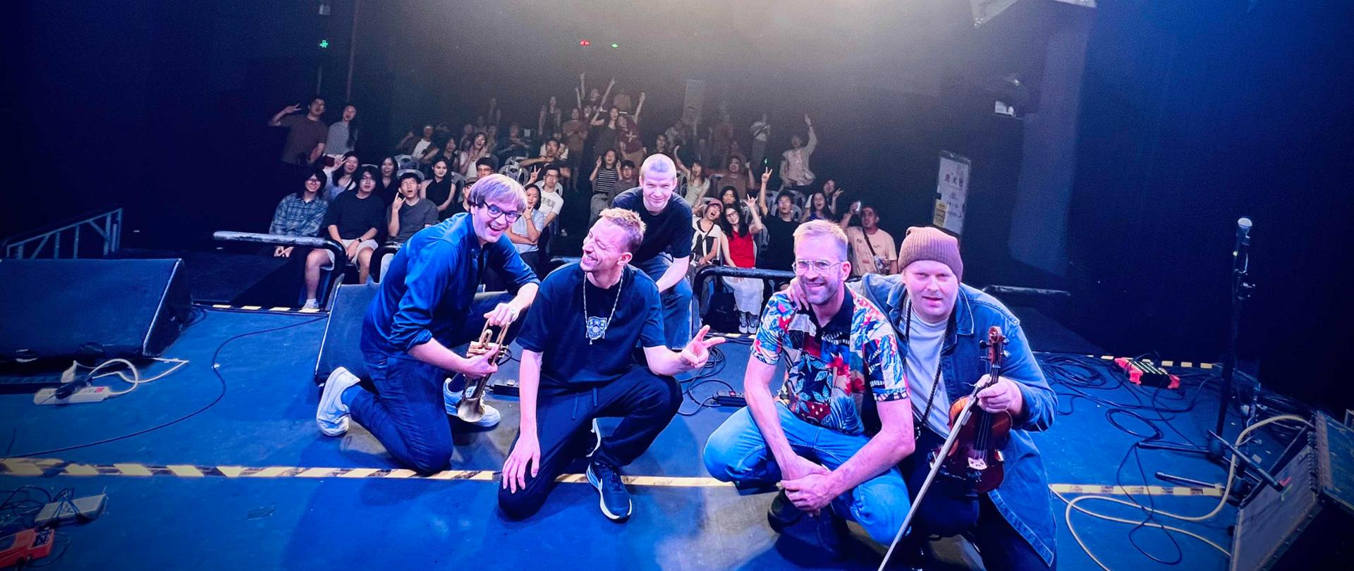 Muzycy i publiczność siedzą razem na scenie po koncercie, ujęcie grupowe w jasnym świetle reflektorów.