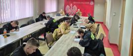 Strażacy OSP siedzą w ławkach i piszą test na świetlicy, w tle komisja strażacka za nimi czerwne logo PSP