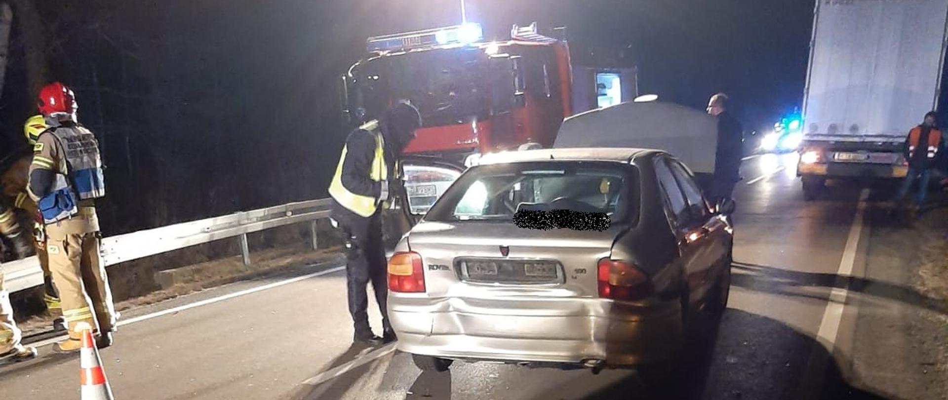Na zdjęciu widać samochód osobowy z uszkodzonym tyłem i podniesioną maską, obok samochodu stoją Strażacy i Policjant. Za samochodem osobowym znajduje się samochód straży pożarnej który oświetla teren działań. Po prawej stronie widać tył naczepy samochodu ciężarowego stojącego na poboczu.