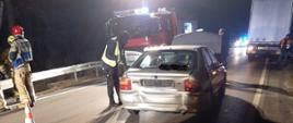 Na zdjęciu widać samochód osobowy z uszkodzonym tyłem i podniesioną maską, obok samochodu stoją Strażacy i Policjant. Za samochodem osobowym znajduje się samochód straży pożarnej który oświetla teren działań. Po prawej stronie widać tył naczepy samochodu ciężarowego stojącego na poboczu.