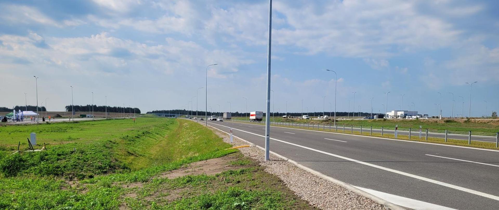 Nowoczesna, wielopasmowa droga przebiegająca przez otwarty teren. Po lewej stronie widoczny jest pas zieleni i rów odwadniający, a po prawej utwardzone pobocze z kruszywa. Wzdłuż jezdni ustawiono wysokie latarnie uliczne. Na drodze porusza się kilka samochodów i ciężarówek. W tle znajdują się niskie zabudowania przemysłowe oraz pas lasu. Nad krajobrazem rozciąga się niebo z dużą liczbą jasnych, rozproszonych chmur.