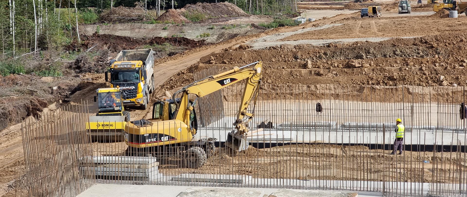 Budowa drogi ekspresowej. na pierwszym planie fundamenty obiektu mostowego, w oddali pracujący sprzęt budowlany.