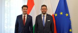Przed białą ścianą stoją wiceminister Szeptycki i mężczyzna w ciemnogranatowym garniturze, za nimi flagi Polski, Węgier i UE, po lewej zasłonięte firankami okno.
