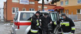 Zdjęcie przedstawia samochód strażacki na tle budynku Centrum Medycznego w Wilczycach. Na zdjęciu widać jak dwóch strażaków pomaga wysiąść starszemu mężczyźnie z samochodu, który wyraził chęć zaszczepienia się przeciwko COVID-19. 
