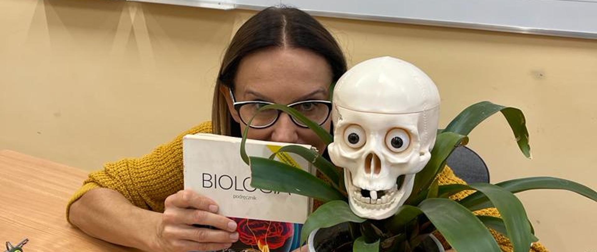 Zdjęcie nauczycielki biologii p. Haliny Pospieszałowskiej, która ukrywa się za podręcznikiem do biologii. Obok na biurku kwiatek w doniczce. Za plecami tablica szkolna.