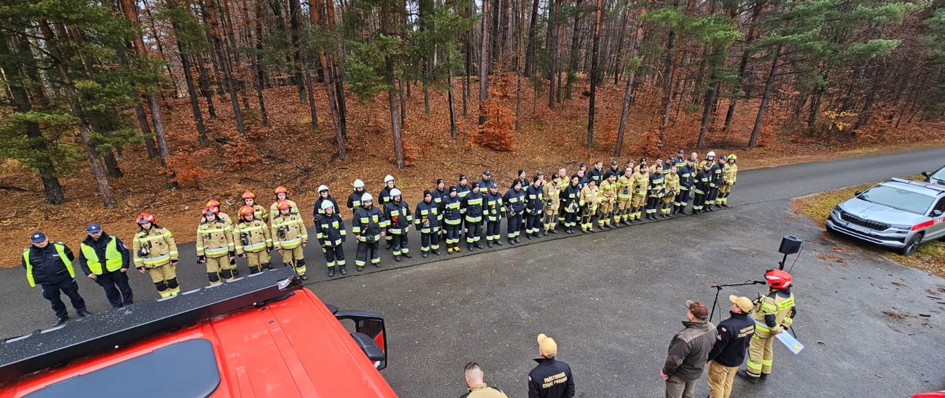 Zdjęcie przedstawia strażaków na zbiórce, w tle widoczny las. 