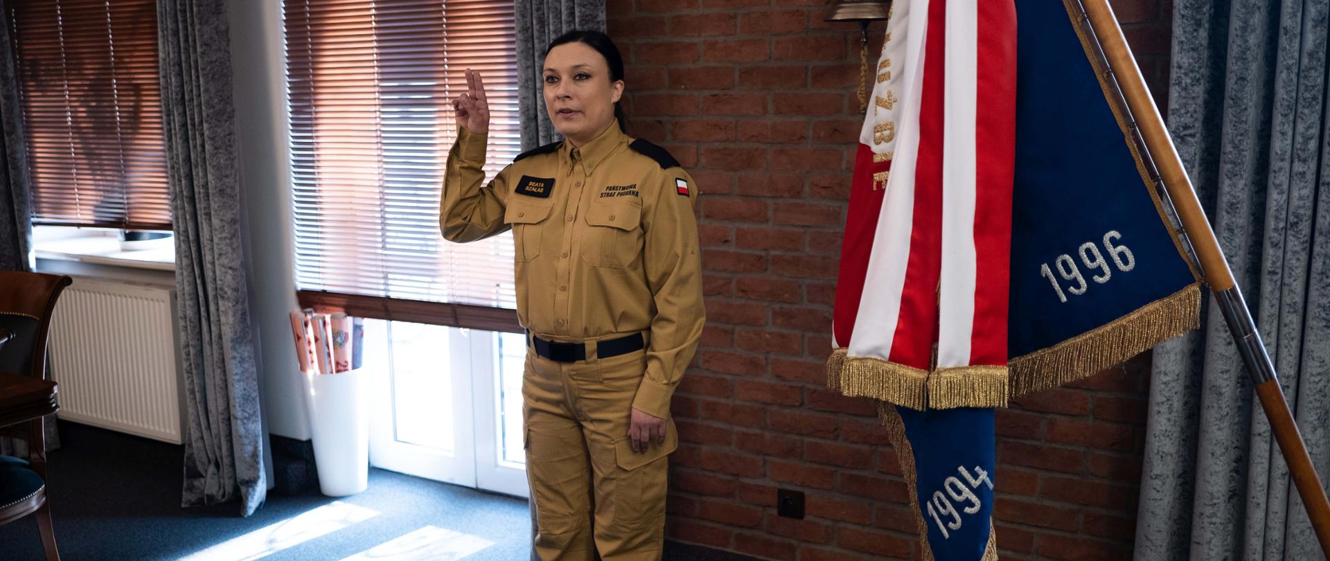 Gabinet Komendanta Centralnej Szkoły PSP, stojąca na baczność kobieta strażak w piaskowym mundurze z uniesioną prawą dłonią w chwili składania uroczystego ślubowania, po prawej widoczny Sztandar Szkoły