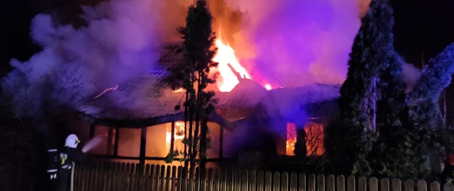 Pożar drewnianego budynku mieszkalnego jednorodzinnego. Budynek całkowicie objęty pożarem. Przed budynkiem strażacy prowadzący działania gaśnicze.