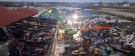 pogorzelisko i zerwany dach w hali produkcyjnej widoczne pojemniki z chemikaliami częściowe pokrycie hali czerwoną blachą falistą duża ilość materiałów izolacyjnych rozrzucona dookoła, koparka przeładunkowa w tle dwa pojazdy straży pożarnej woda z pianą na terenie placu