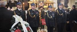 Strażacy w mundurach galowych stoją na baczność przed trumną śp. płk. poż. Stanisława Luli. Obok widoczny wieniec z biało-czarną wstęgą pożegnalną oraz medalami i odznaczeniami zasłużonego oficera.