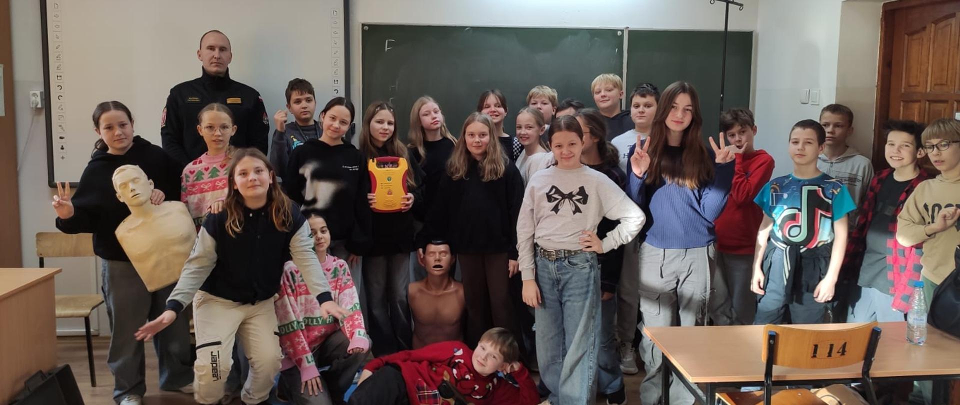 Sala lekcyjna. Pod tablicą do zdjęcia pozują dzieci i strażak.
