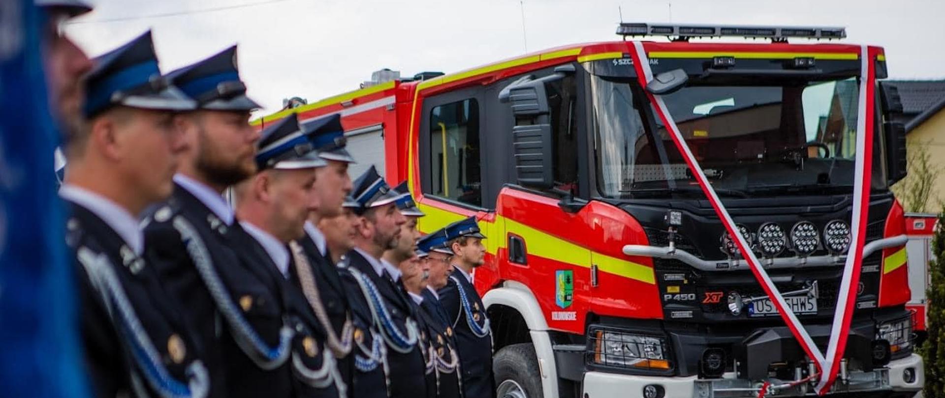 Na zdjęciu znajdują się strażacy ustawieni w szeregu a w tel stoi samochód strażacki