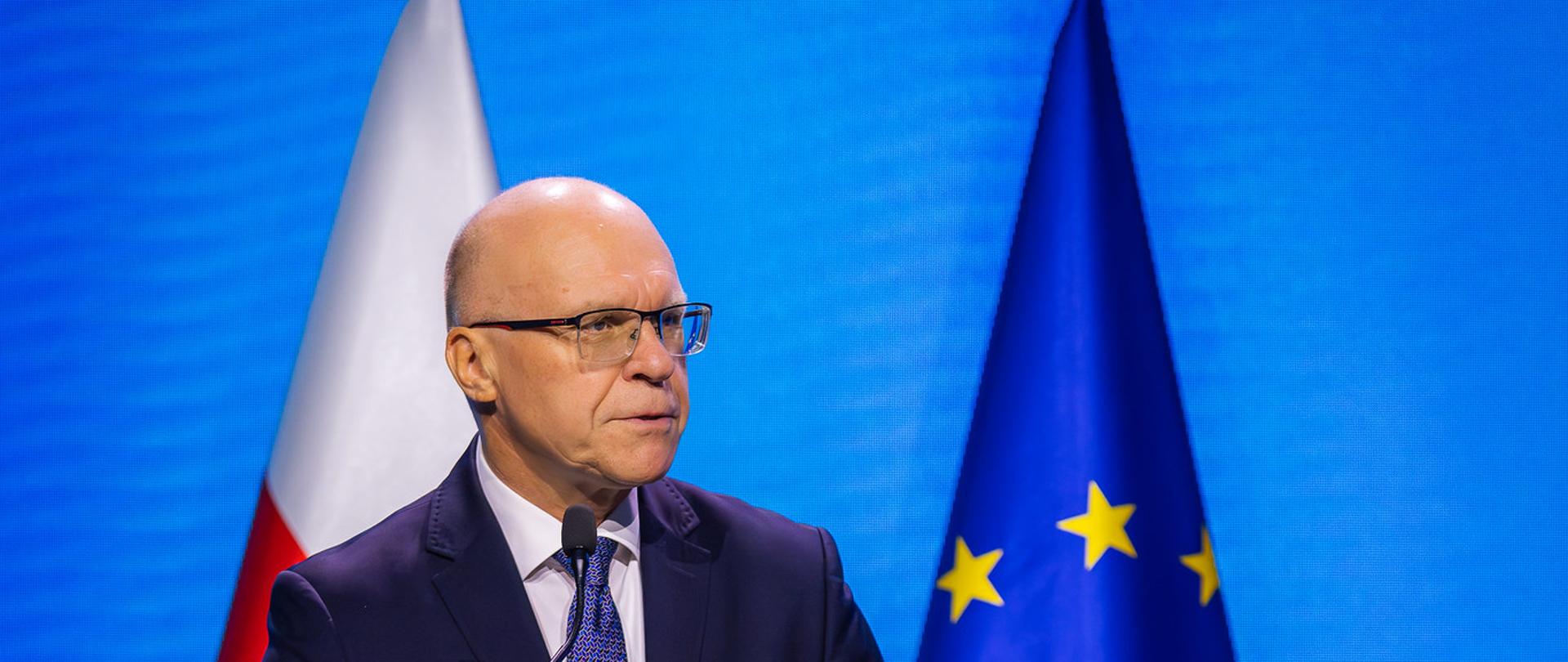 Ambasador Rafał Wiśniewski stojący przy podium na tle flag. 