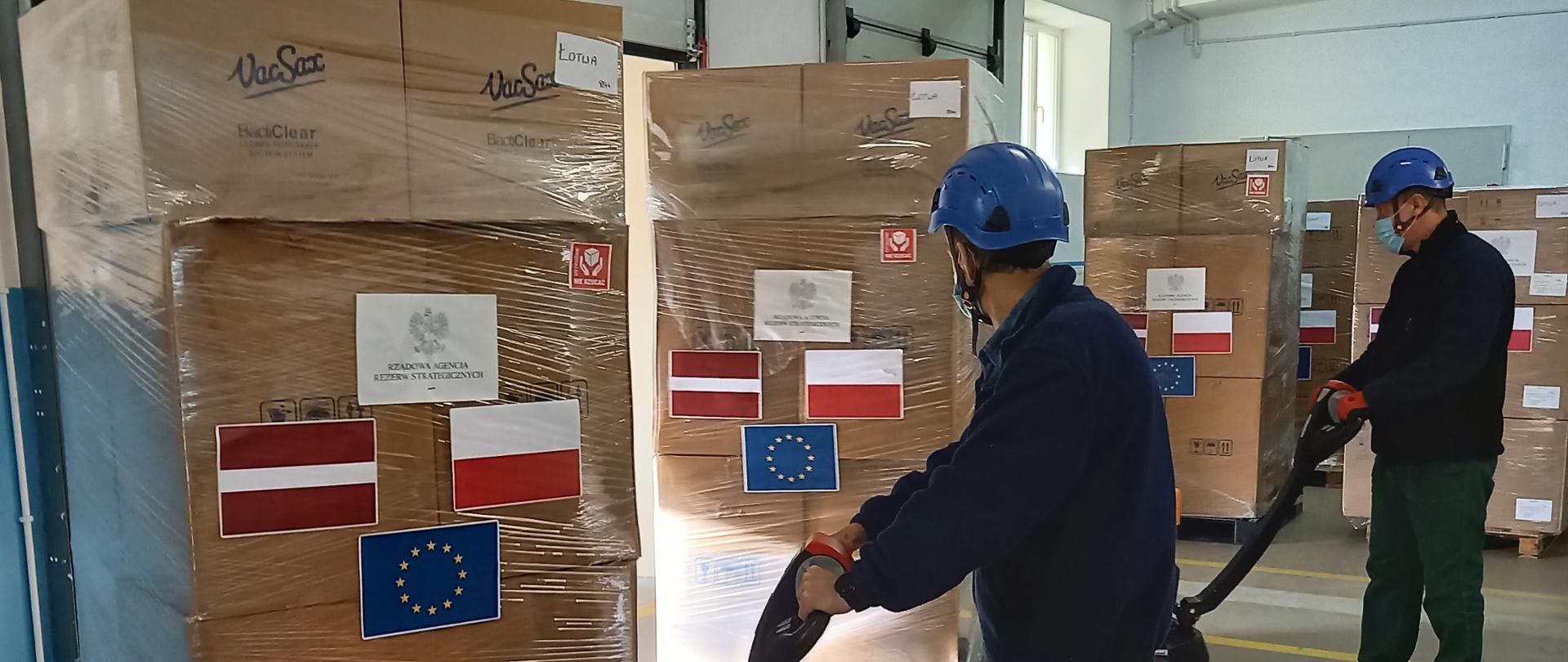 Dwóch mężczyzn wiezie na wózkach widłowych palety, na których ustawione są zapakowane w folię duże pudełka. Na na paczkach naklejone są flagi Łotwy, Polski i Unii Europejskiej 