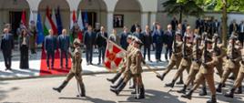 Na pierwszym planie defilada wojskowa. Na czerwonym dywanie stoją premierzy: Donald Tusk i Péter Magyar. Po lewej i prawej stronie premierów stoją członkowie rządu Polski i Węgier. W tle 9 flag (3 flagi Polski, 3 Węgier i 3 UE). Przy flagach stoi 6 żołnierzy (dwie trójki). W tle fasada budynku z tarasem oraz inne osoby. Po prawej stronie drzewa.