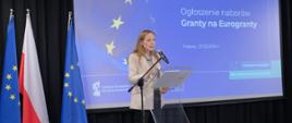 Minister funduszy i polityki regionalnej Katarzyna Pełczyńska-Nałęcz stoi na scenie przy pulpicie z mikrofonem, po lewej stronie flagi Polski i UE, za plecami pani minister grafika na niebieskim tle z informacją o ogłoszeniu naborów