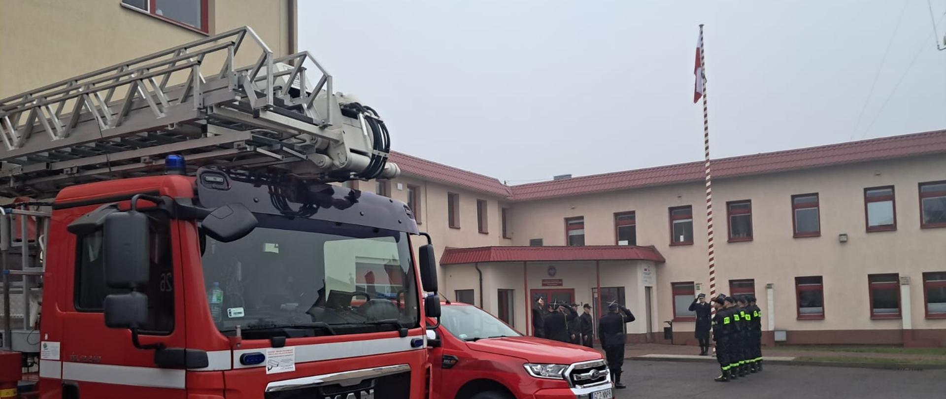 Uroczysta zmiana służby z okazji Narodowego Święta Niepodległości. Strażacy ze zmian służbowych stoją naprzeciwko siebie. Poczet flagowy wciąga flagę państwową na maszt. Komendant, dowódca jednostki, poczet sztandarowy i dowódcy zmian salutują. 