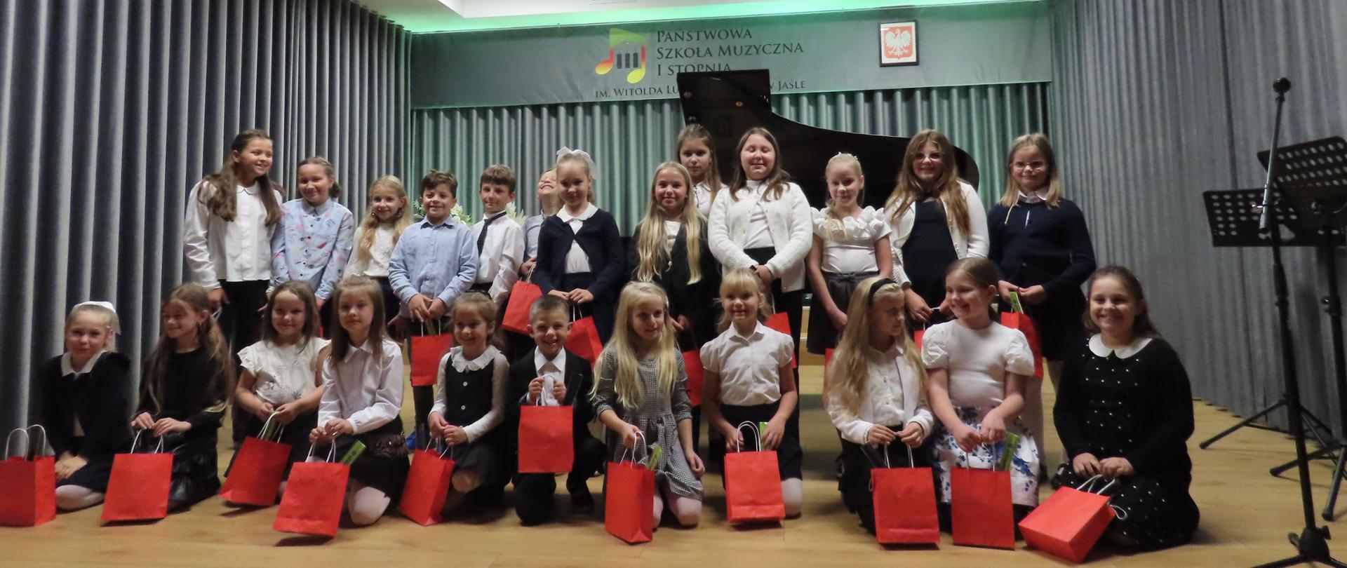Na zdjęciu widzimy pierwszoklasistów Państwowej Szkoły Muzycznej I st. im. Witolda Lutosławskiego w Jaśle po uroczystości pasowania na ucznia. Dzieci w eleganckich strojach stoją na scenie, trzymając upominki, a radosny uśmiech na ich twarzach podkreśla wyjątkowy charakter tego dnia.