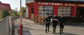 Kolorowa fotografia wykonana w pogodny dzień na zewnątrz budynku Komendy Powiatowej PSP w Brzozowie. Na pierwszym planie trzech strażaków z flagą udają się w kierunku masztu flagowego. W tle znajdują się strażacy stojący w szeregu, część z nich ubrana w ubrania służbowe, a część w koszarowe.