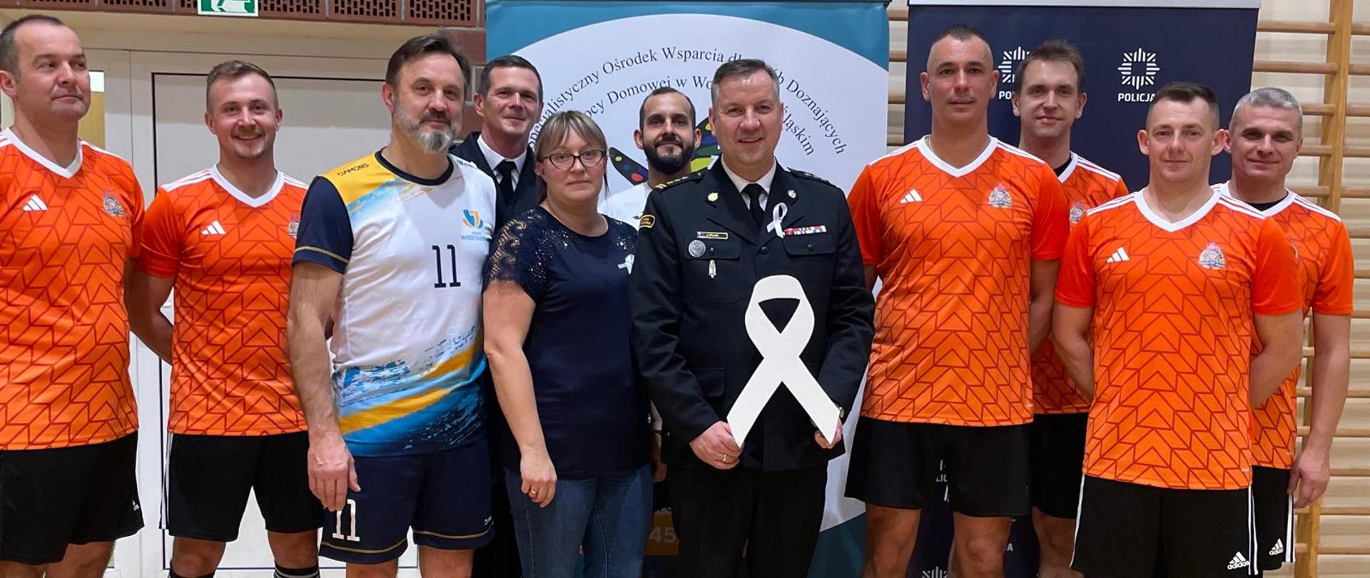 Uczestnicy turnieju siatkówki w ramach kampanii Biała Wstążka – strażacy, policjanci i pracownicy starostwa – pozują wspólnie w sali gimnastycznej. Pośrodku stoi funkcjonariusz trzymający dużą białą wstążkę, symbol sprzeciwu wobec przemocy