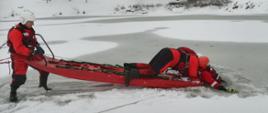 zdjęcie przedstawia zamarznięty zbiornik wodny i dwóch strażaków, którzy przy pomocy sań lodowych wyciągają poszkodowanego strażaka z wody.