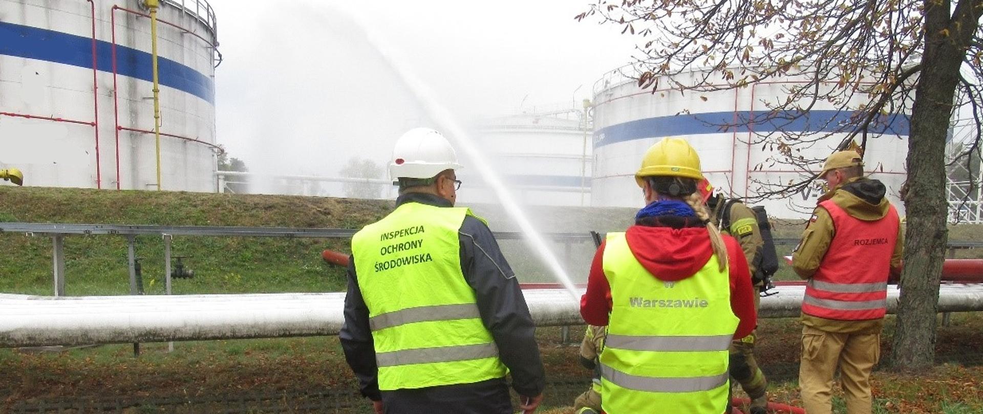 Inspektorzy Wojewódzkiego Inspektoratu Ochrony Środowiska w Warszawie stoją na otwartym terenie i monitorują akcję gaśniczą prowadzoną przez funkcjonariuszy Państwowej Straży Pożarnej. W tle widać kilkunastometrowej wysokości białe zbiorniki o pojemności 5000 m3.