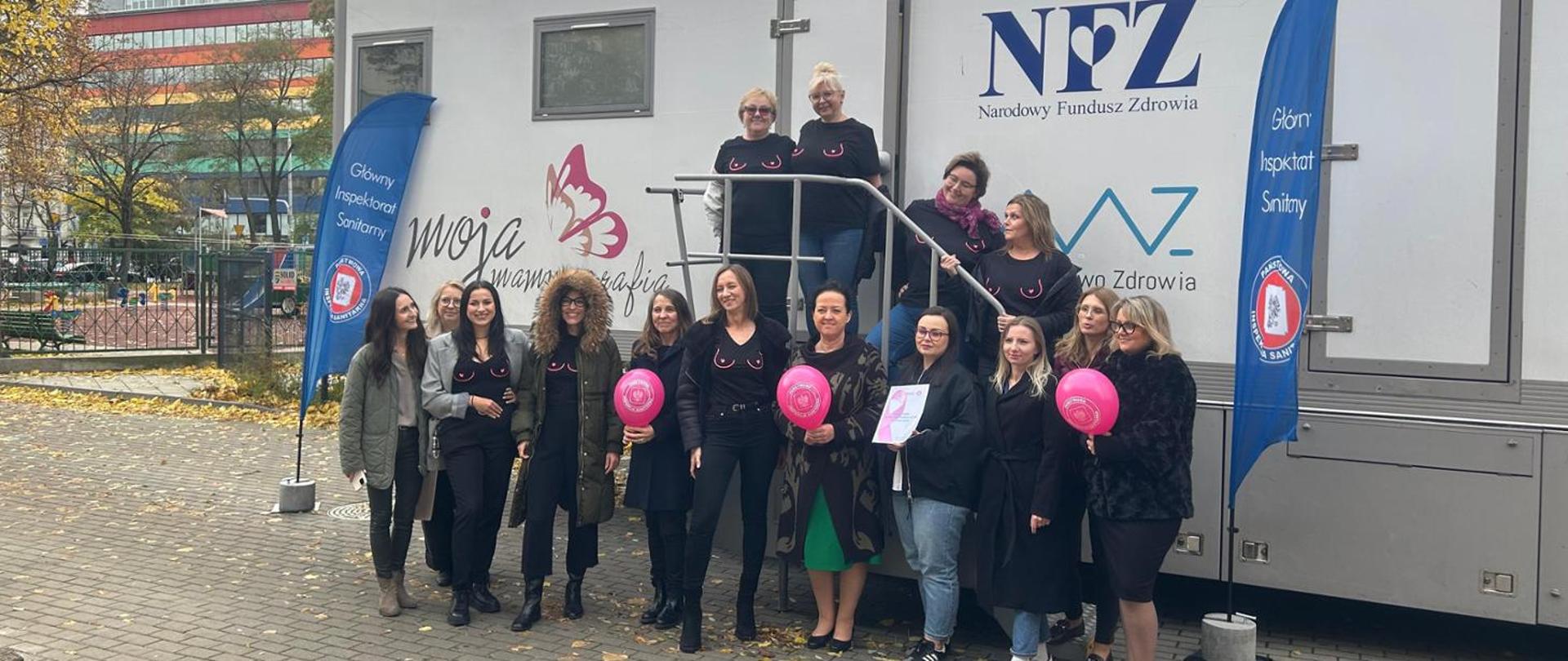 Na zdjęciu widać grupę kobiet stojących przed mobilnym punktem medycznym z logo NFZ (Narodowy Fundusz Zdrowia) oraz napisami „Moja mammografia” i „Główny Inspektorat Sanitarny”.
Kobiety uśmiechają się i pozują do wspólnego zdjęcia. Część z nich ma na sobie czarne koszulki z różowym motywem graficznym przypominającym zarys biustu – symbol kampanii profilaktycznej dotyczącej badań piersi. Niektóre trzymają różowe balony lub materiały promocyjne.
Zdjęcie wykonano na zewnątrz, w jesiennej scenerii – widać drzewa z żółtymi liśćmi i kolorowy budynek w tle. Całość ma charakter akcji zdrowotnej promującej profilaktykę raka piersi i zachęcającej kobiety do badań mammograficznych.