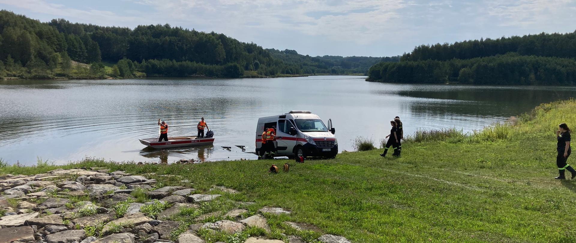 Na zdjęciu widoczna łódź płaskodenna zwodowana na akwenie Korzeń, na której znajduje się dwóch ratowników zabezpieczonych w kamizelki ratunkowe. Obok znajduje się samochód SLRw OSP KRSG Skrzyszów wraz z przyczepą do łodzi stojącą w wodzie. 