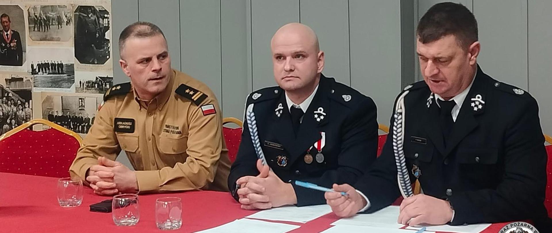 Trzech strażaków siedzących za stołem prezydialnym w trackie trwania spotkania sprawozdawczo-wyborczego w OSP. Wśród nich jest komendant miejski PSP w Łomży.
