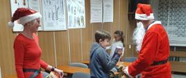 Mężczyzna w stroju Mikołaja i kobieta w czerwonym stroju elfa rozdają dają chłopcu stojącemu na środku sali lekcyjnej pomiędzy ławkami czekoladę. Z tyłu stoi dziewczynka.