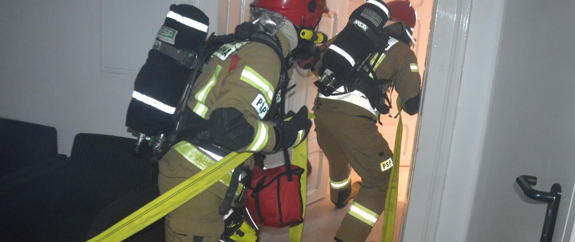 Ćwiczenia w Domu Kultury w Rawiczu. Wnętrze budynku. Dwóch strażaków z linia gaśniczą przeszukuje pomieszczenia obiektu w celu zlokalizowania źródła ognia. Poruszają się jeden za drugim. Strażak znajdujący się z tyłu przez ramię ma przewieszoną torbę z linką ratowniczą, a na szyi zawieszoną kamerę termowizyjną. 