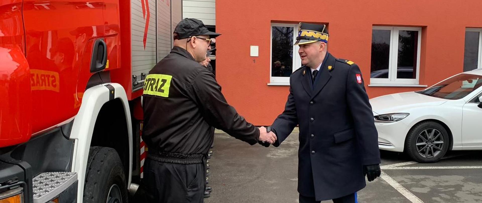 strażak w rogatywce i płaszczu wita się ze strażakiem w czapeczce i czarnym mundurze, w tle czerwony samochód strażaki oraz biały osobowy, za nimi budynek 