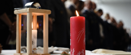 Światełko betlejemskie oraz czerwona świeca stoją na stole obok leżą opłatki i stoją policjanci oraz strażacy.