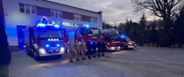 Na zdjęciu widać radomskich strażaków, którzy stoją w postawie zasadniczej na baczność i oddają hołd zmarłemu na służbie strażakowi. Za nimi ustawione są wozy straży pożarnej z włączonymi sygnałami sygnalizacyjno dźwiękowymi.