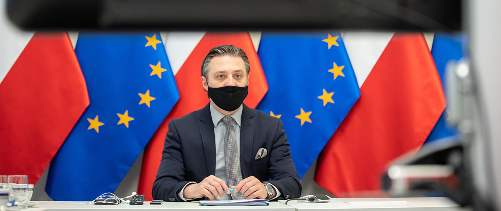Na zdjęciu widać wiceministra Bartosza Grodeckiego siedzącego za stołem na tle flag Polski i UE wpatrującego się w ekran.