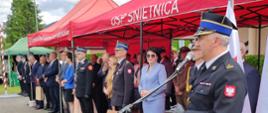 Na zdjęciu przy mównicy na uroczystości 75-lecia OSP Uście Gorlickie oraz poświęceniu i przekazaniu samochodów pożarniczych przemawia zastępca komendanta powiatowego. W tle czerwone namioty i zaproszeni na uroczystość goście mundurowi i cywile.
