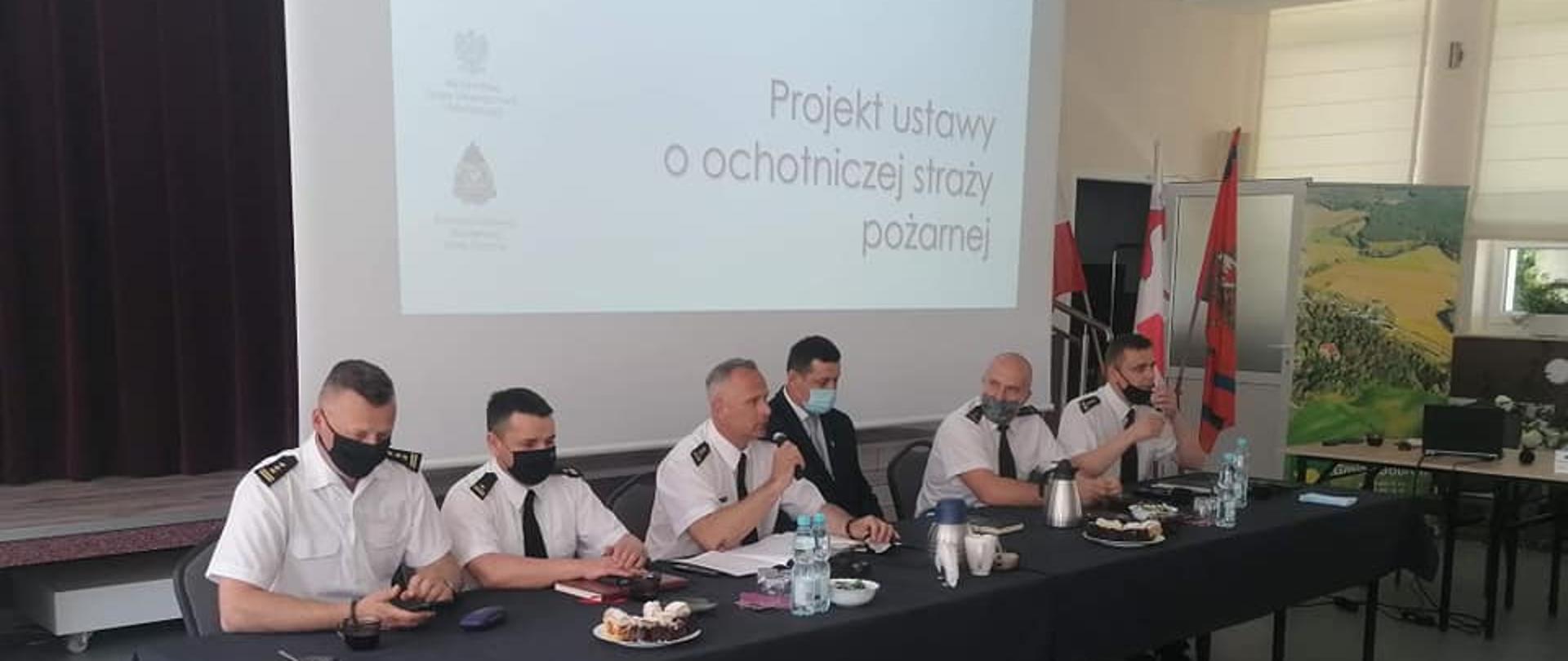 NA ZDJĘCIU WIDAĆ 6 MĘŻCZYZN SIEDZĄCYCH ZA DŁUGIM PROSTOKĄTNYM STOŁEM , NAKRYTYM CZARNYM OBRUSEM. 5 MĘŻCZYZN MA NA SOBIE BIAŁE KOSZULE SĄ TO STRAŻACY , 1 MĘŻCZYZNA MA NA SOBIE CZARNĄ MARYNARKĘ. 