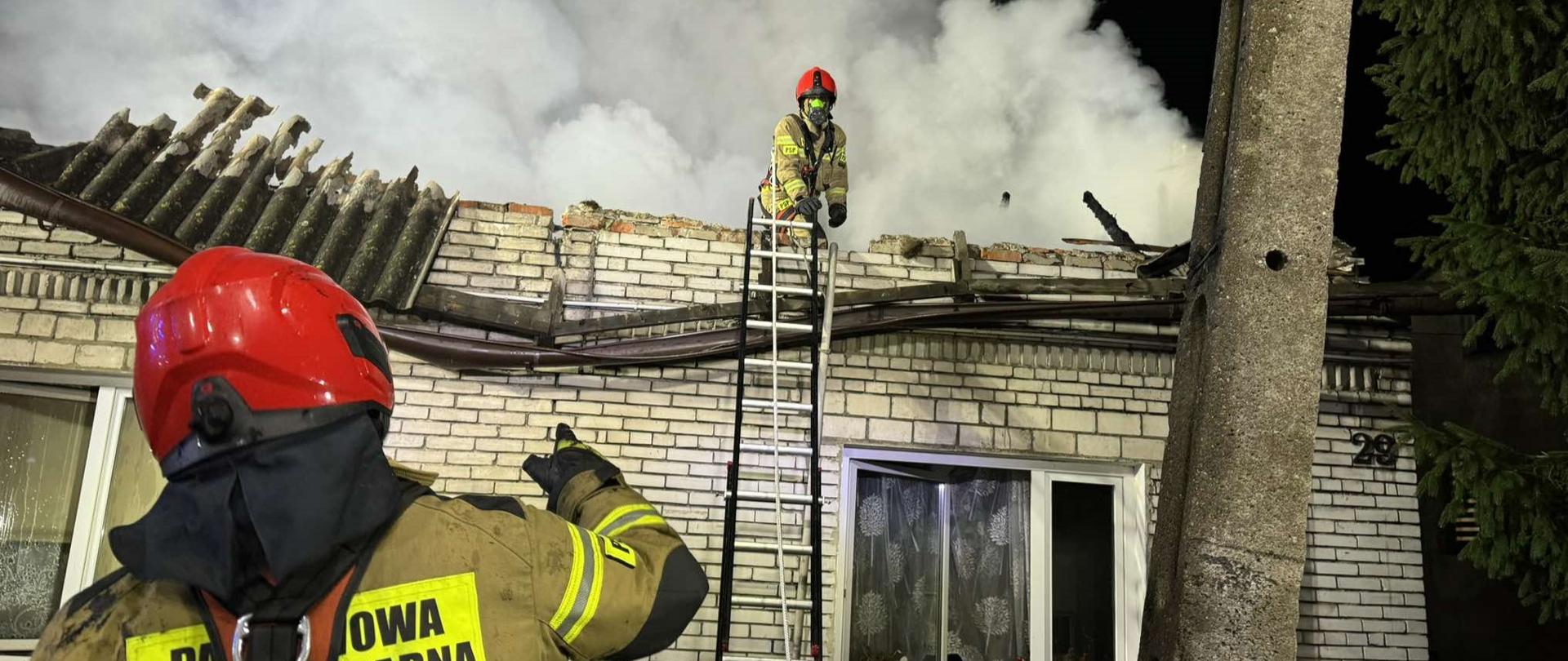 Na zdjęciu widać dwóch strażaków w pełnym umundurowaniu i hełmach bierze udział w akcji gaśniczej. Jeden strażak stoi na dole i wskazuje, a drugi znajduje się na drabinie, na wysokości dachu, z którego unosi się gęsty dym. Dach jest spalony. W tle widoczny jest budynek z cegły z oknami.