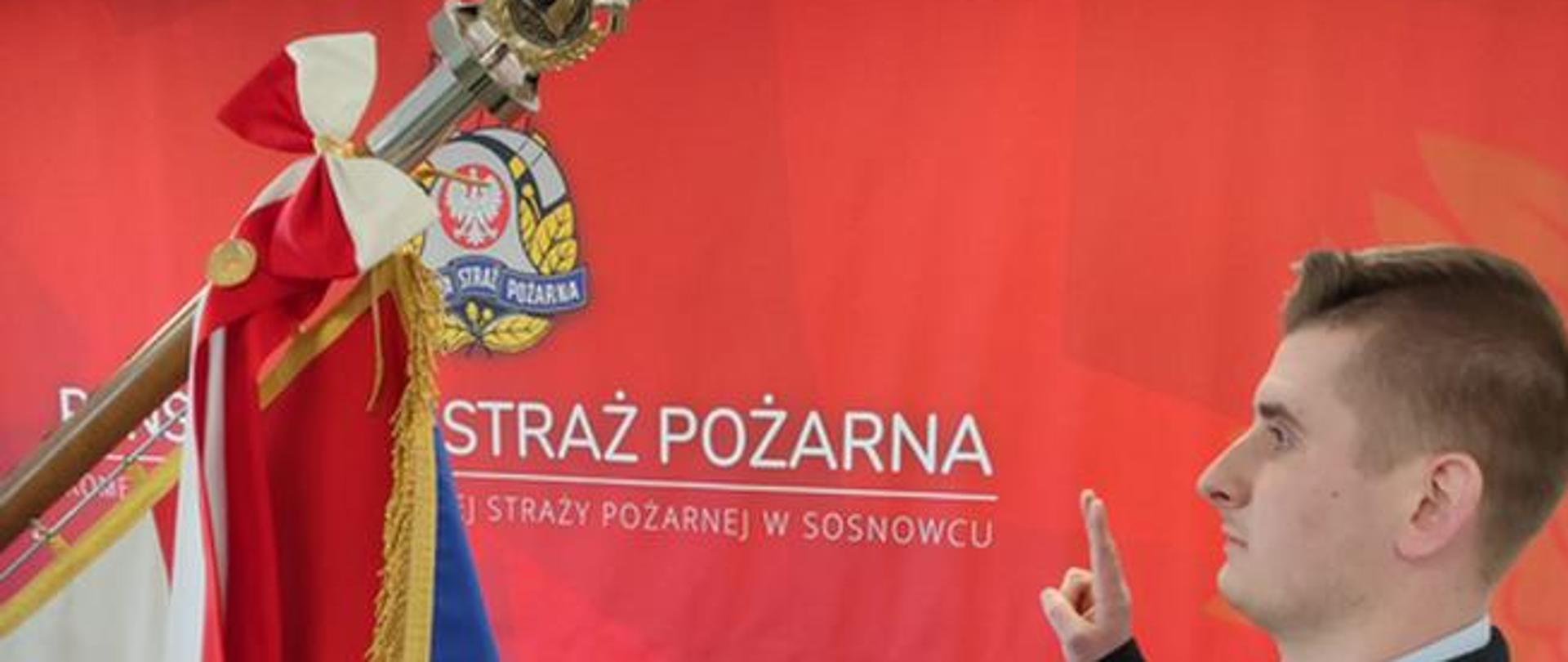 Po prawej stronie zdjęcia ślubujący strażak stojący naprzeciwko sztandaru. Po lewej stronie zdjęcia widoczny poczet sztandarowy Komendy Miejskiej PSP w Sosnowcu.