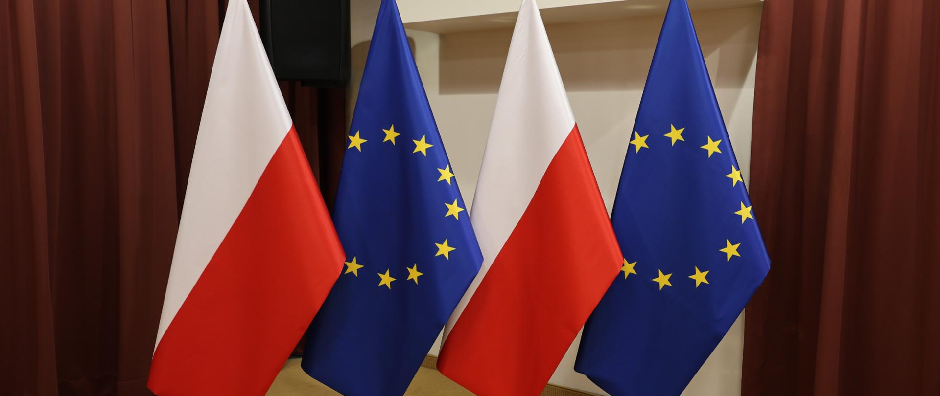 Flagi Polski i Unii Europejskiej na tle kremowej ściany oraz bordowych kotar. 