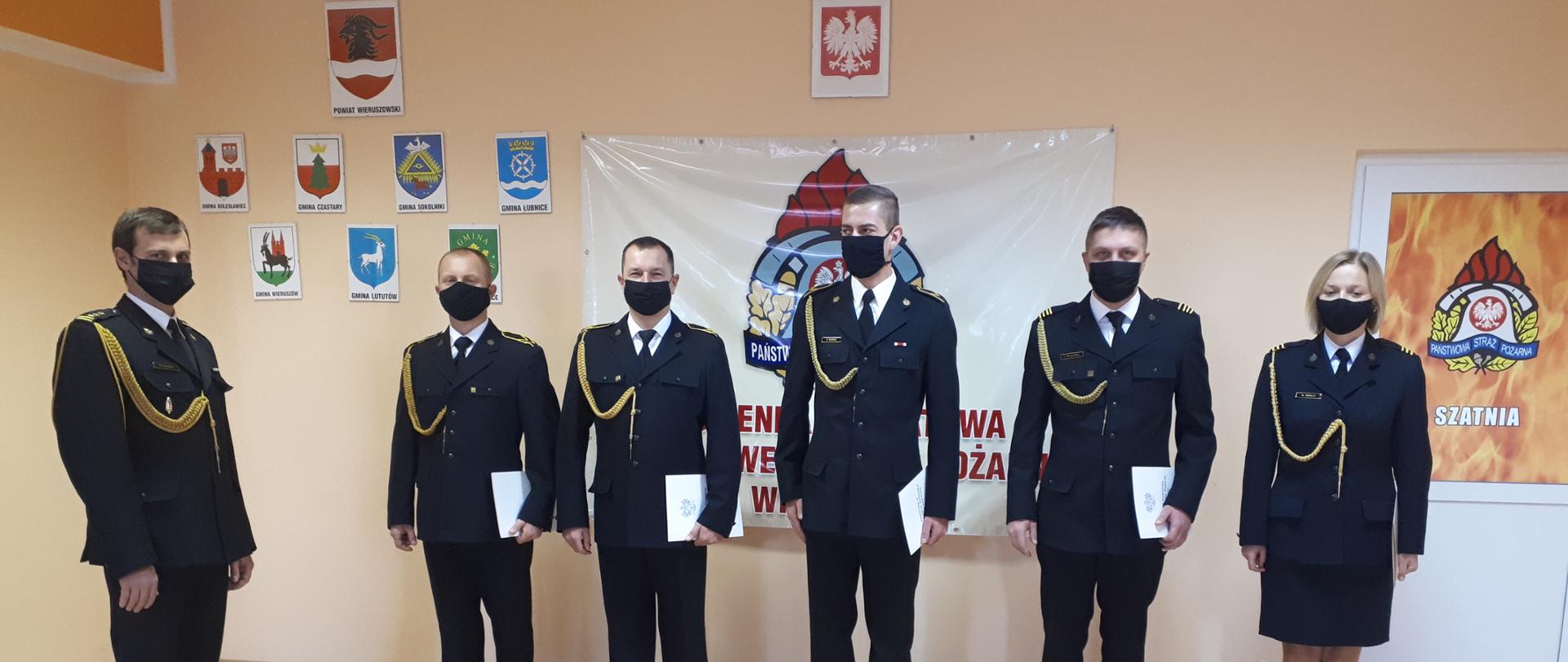 Zdjęcie przedstawia osoby awansowane na wyższe stopnie służbowe ustawione w szeregu obok siebie w sali szkoleniowej tutejszej jednostki ratowniczo-gaśniczej. W tle znajdują się herb powiatu oraz herby wszystkich gmin powiatu wieruszowskiego. Funkcjonariusze ubrani są w mundury wyjściowe wraz ze sznurami galowymi z założonymi na twarz maseczkami ochronnymi. Od lewej strony bokiem ustawiony stoi komendant powiatowy, następnie w odstępach stoi czterech awansowanych funkcjonariuszy. Ostatnią osobą w szeregu jest funkcjonariuszka.