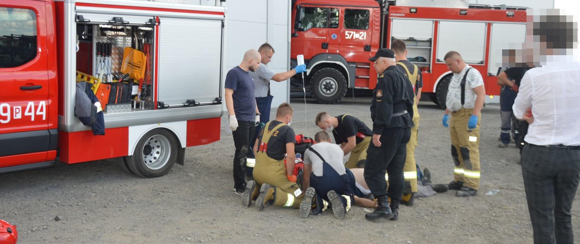 Teren jednej z firm w Kołaczkowicach. Przed halą strażacy udzielający kwalifikowanej pierwszej pomocy mężczyźnie, u którego stwierdzono NZK. Część z nich stoi, pozostali klęczą przy poszkodowanym. Jeden ze strażaków trzyma kroplówkę. Widoczne są również dwa wozy strażackie.
