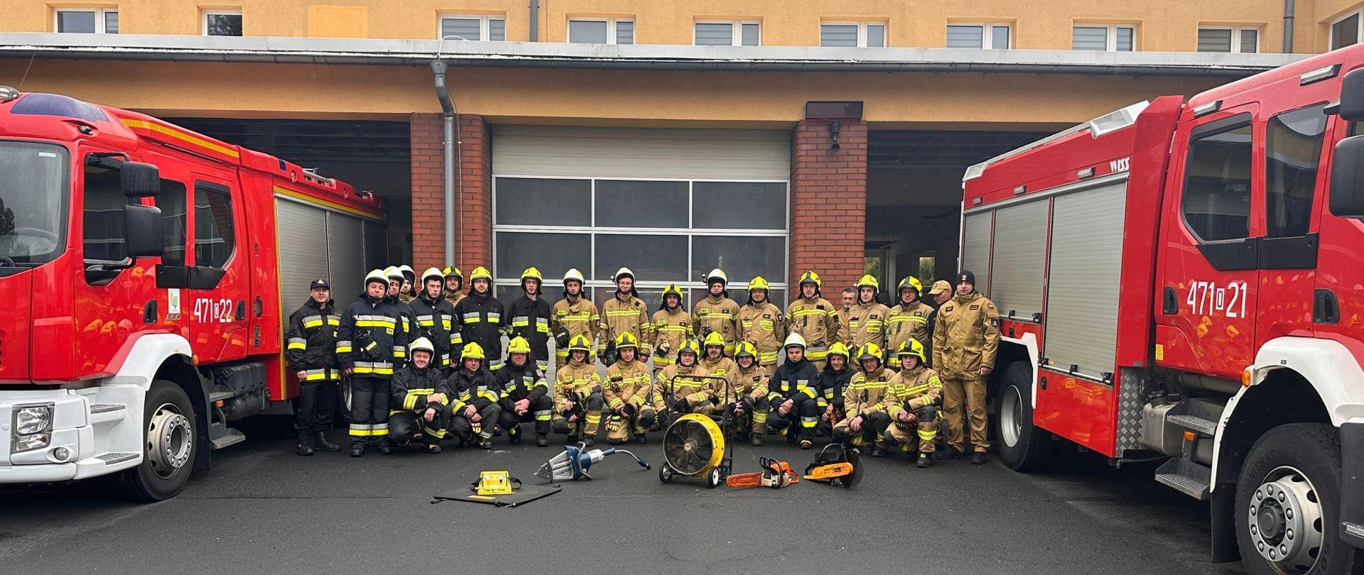 Na tle budynku JRG Olesno, pomiędzy samochodami pożarniczymi ustawieni są uczestnicy kursu wraz z komisją egzaminacyjną. Przed strażakami ustawiony jest sprzęt ratowniczy.
