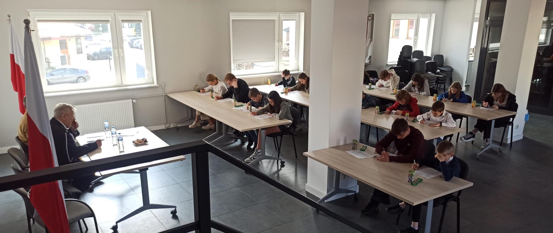 Na pierwszym planie dzieci siedzące przy stołach. Po lewej siedzą członkowie komisji turnieju. Trwa część pisemna turnieju wiedzy pożarniczej.