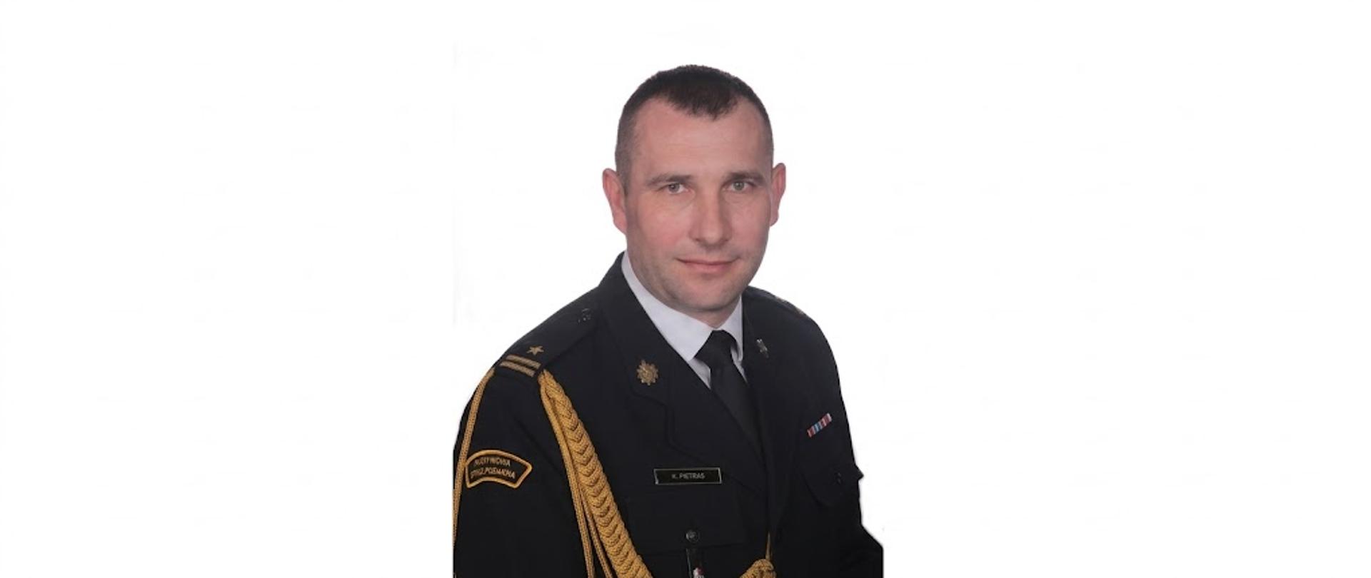 Portret mężczyzny w ciemnym mundurze Państwowej Straży Pożarnej, przedstawiony w bardzo szerokim formacie panoramicznym na białym tle. Mężczyzna ma krótkie, ciemne włosy i patrzy prosto w obiektyw z lekkim uśmiechem. Na lewym ramieniu widoczna jest naszywka z napisem „PAŃSTWOWA STRAŻ POŻARNA” oraz złoty sznur galowy. Na piersi przypięta jest tabliczka identyfikacyjna z nazwiskiem „K. PIETRAS” oraz baretki odznaczeń. Postać jest wycentrowana, a po obu jej stronach znajduje się duża, pusta biała przestrzeń.