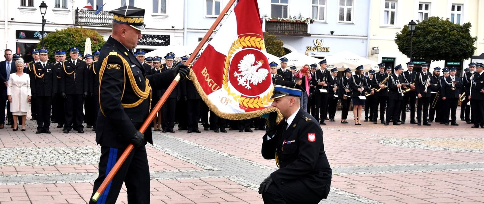 Zdjęcie zrobione na zewnątrz, podczas uroczystości. Oficer Państwowej Straży Pożarnej w stopniu generała trzyma pochylony sztandar. Przed generałem oraz sztandarem klęczy oficer Państwowej Straży Pożarnej w stopniu brygadiera całując materiał sztandaru.
